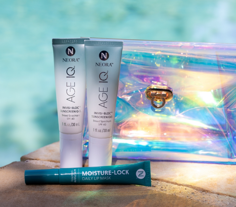 Neora’s Summer Skin Essentials Set, which includes two Age IQ Invisi-Bloc SPF40 Sunscreen Gels and a Moisture-Lock Lip Mask in front of a FREE Holographic Travel Bag laying next to a pool.